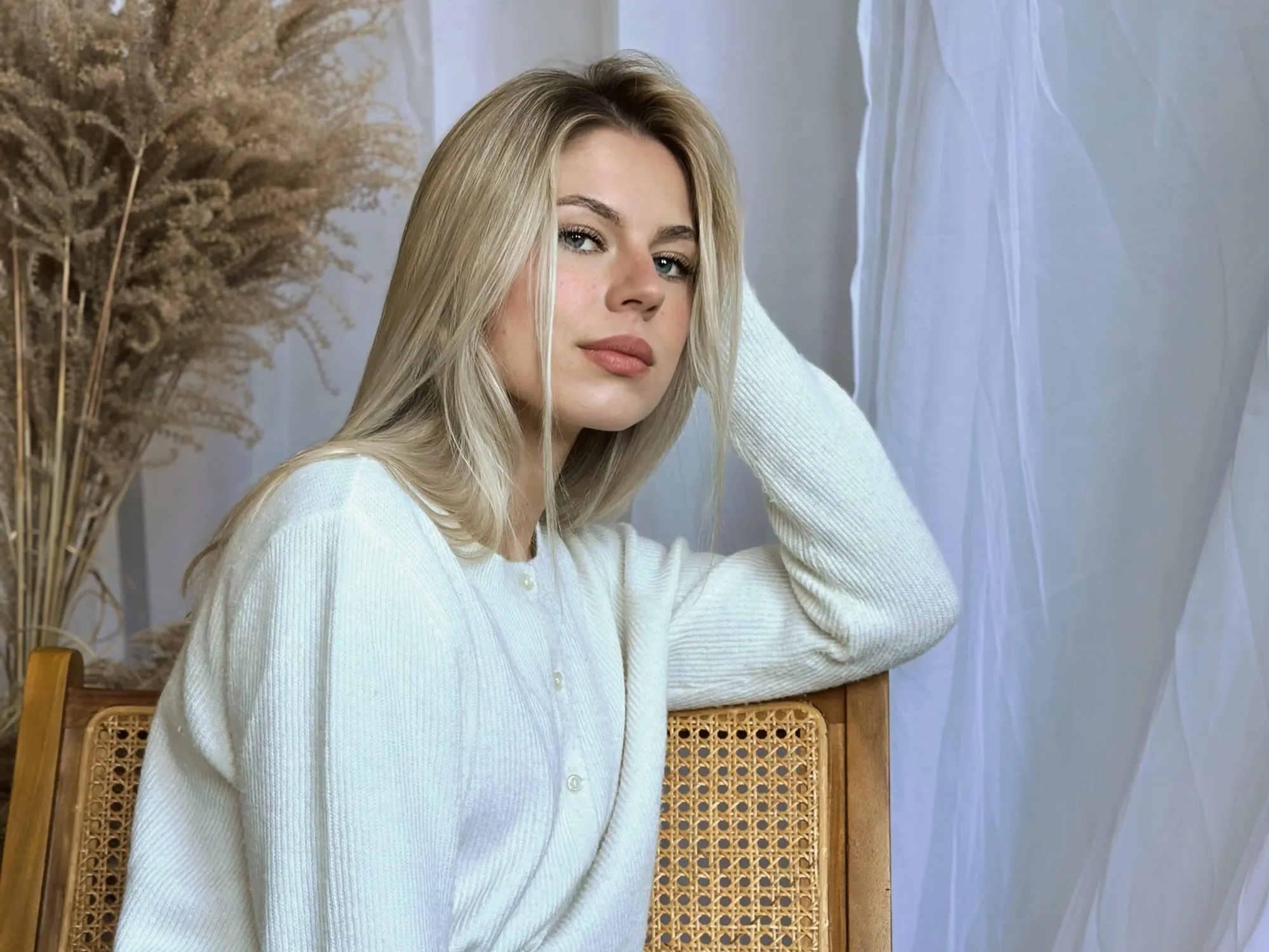 Portret blondynki w białym swetrze na tle jasnej zasłony
